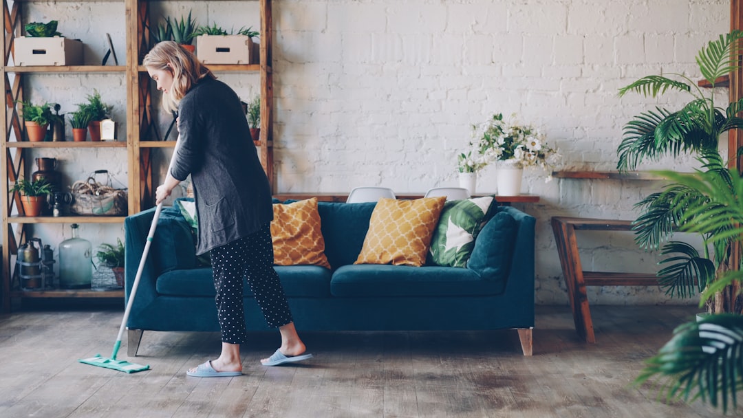 services-01 Attractive young woman is cleaning living room mopping the floor doing housework. Beautiful loft style apartment with modern furniture and green plants is visible.